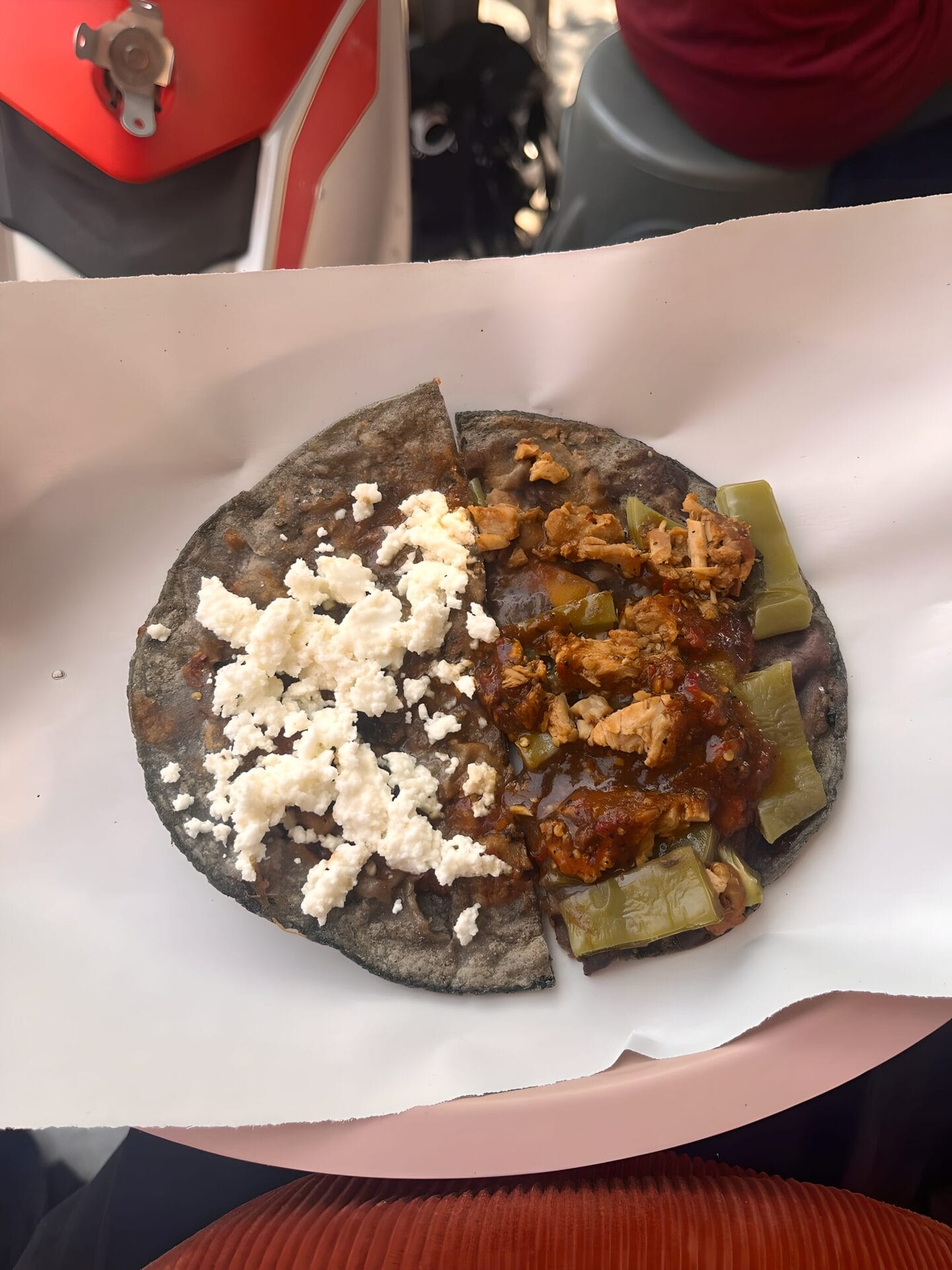 A close-up of a traditional Oaxacan tlayudas in Oaxaca City, Mexico, served on a blue corn tortilla with fresh cheese on one side and a mixture of chicken, green vegetables, and red sauce on the other side.