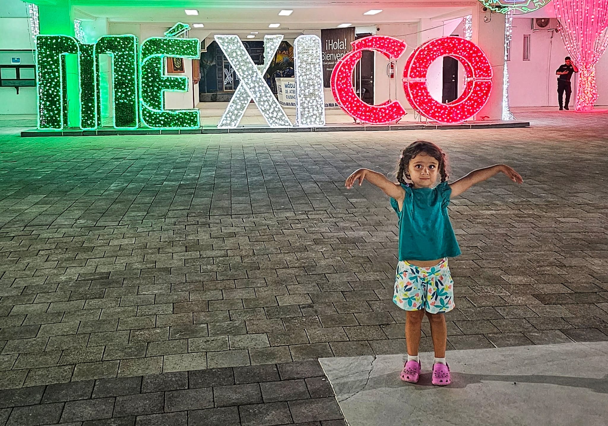 Young girl standing in front of brightly lit "MÉXICO" sign.