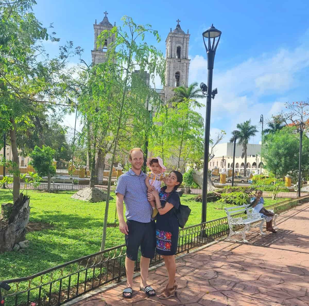 Valladolid Day Trip From Playa Del Carmen A smiling family posing in front of the Cathedral of San Gervasio in Valladolid, Mexico. A father, mother, and their young child are surrounded by lush greenery in a park, with the cathedral's twin towers visible in the background under a clear blue sky.