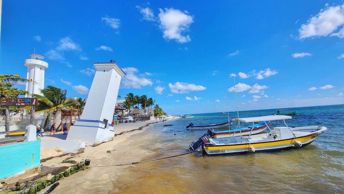 Leaning Lighthouse of Puerto Morelos, Quintana Roo, Mexico, with small boats anchored near the shore on a sunny day.