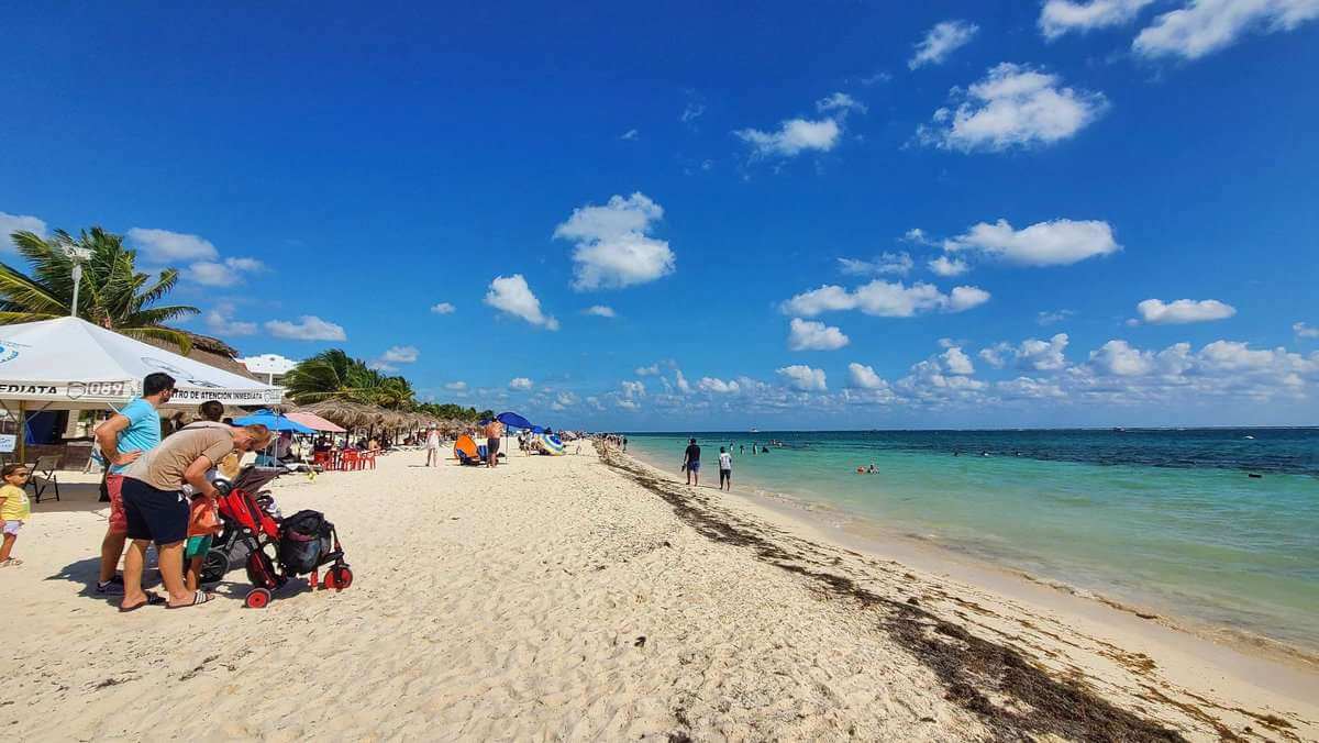 A sunny beach scene at Puerto Morelos, Quintana Roo, Mexico, with people relaxing under tents and umbrellas, and others walking along the shoreline or swimming in the clear blue sea.