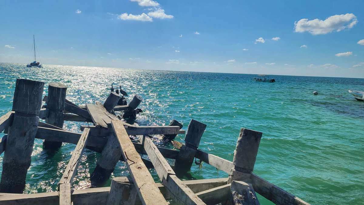 A broken wooden pier extending into the clear, turquoise waters of the Caribbean in Puerto Morelos, Quintana Roo, Mexico. Boats are visible in the distance under a bright blue sky.