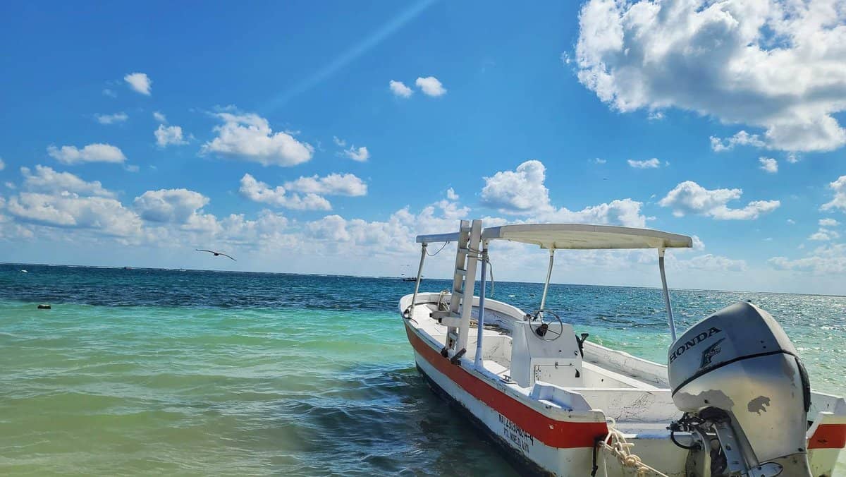 Small boat anchored in the clear blue waters of Puerto Morelos, Quintana Roo, Mexico, with a bright blue sky and scattered clouds in the background.