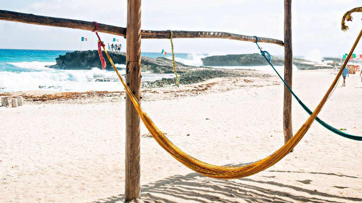 Colorful hammocks hanging under wooden poles on a sandy beach with a rocky shoreline and the turquoise sea in the background in Isla Mujeres, Quintana Roo, Mexico.