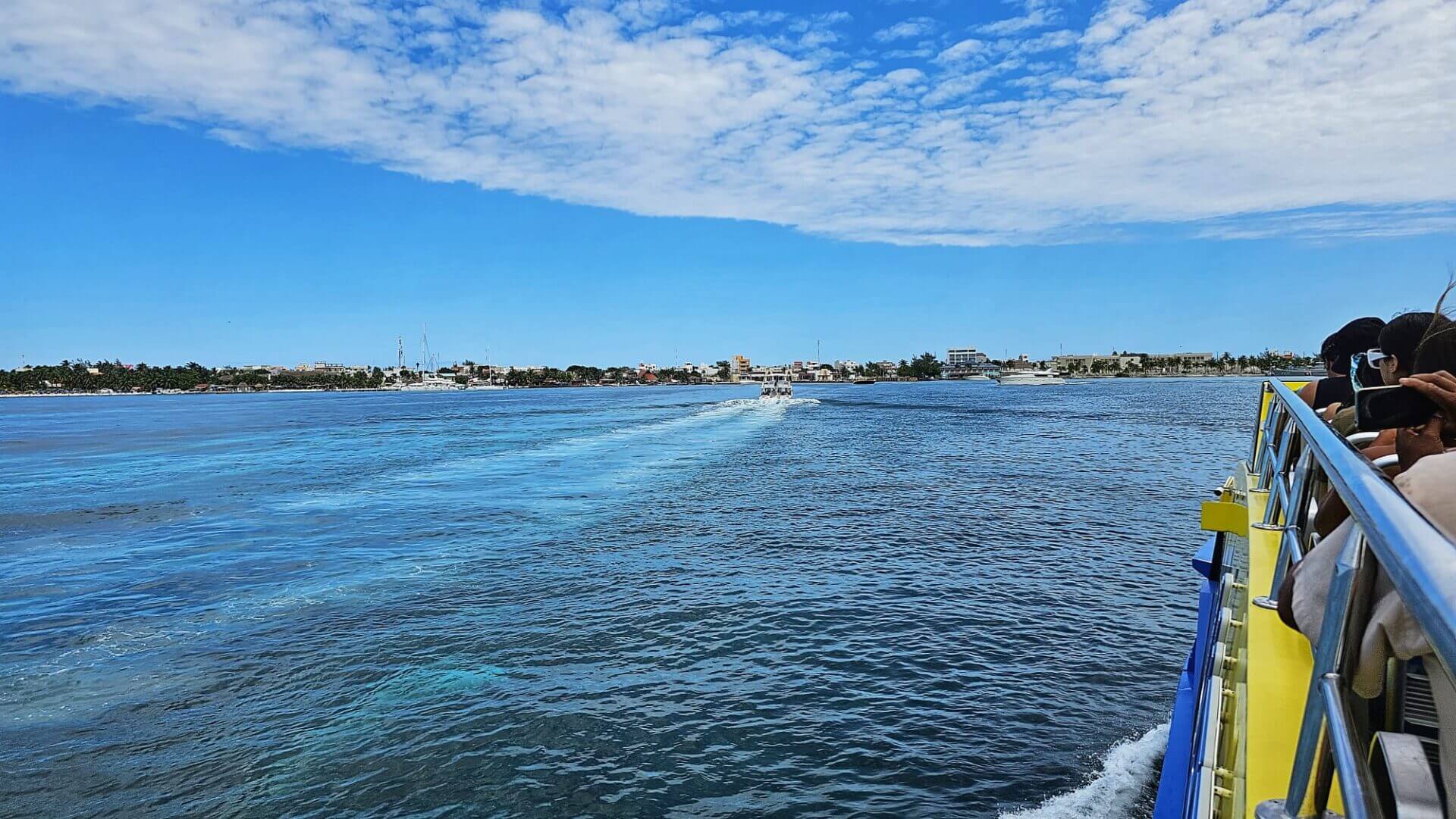 View from a boat of the vibrant waters and distant shoreline of Isla Mujeres, Quintana Roo, Mexico, under a partly cloudy sky.