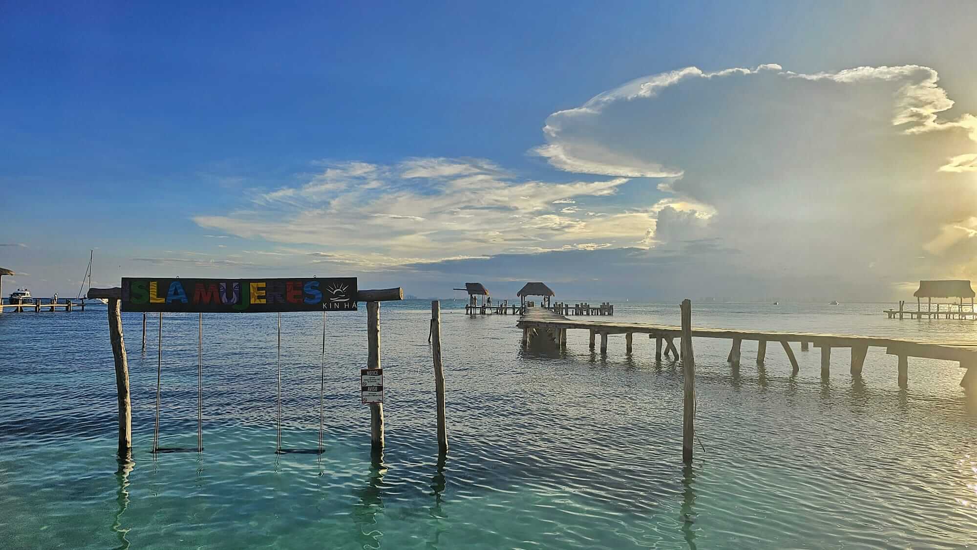 A picturesque view of a turquoise ocean with a wooden pier extending into the water at Isla Mujeres, Quintana Roo, Mexico. The sky is clear with some clouds, and a colorful sign reads "Isla Mujeres" above two swings hanging over the shallow water. In the distance are more piers with huts.
