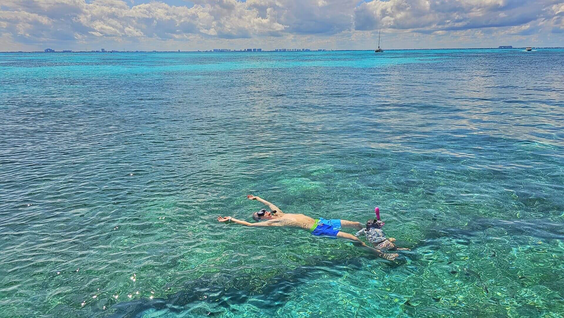Person snorkeling in crystal-clear turquoise waters off the coast of Isla Mujeres, Quintana Roo, Mexico, with a distant view of boats and a city skyline on the horizon under a partly cloudy sky.