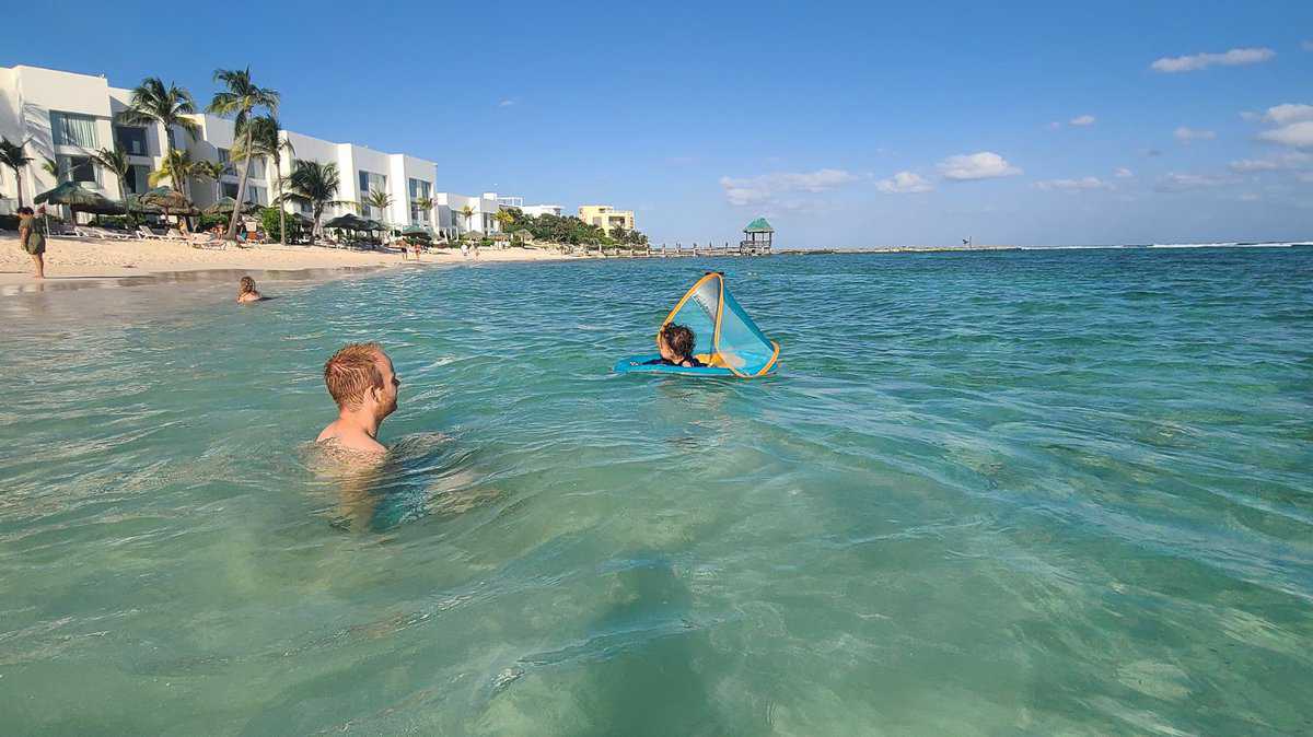 Two people enjoy the clear azure waters in Akumal, Quintana Roo, Mexico with a sandy beach and coastal buildings in the background.