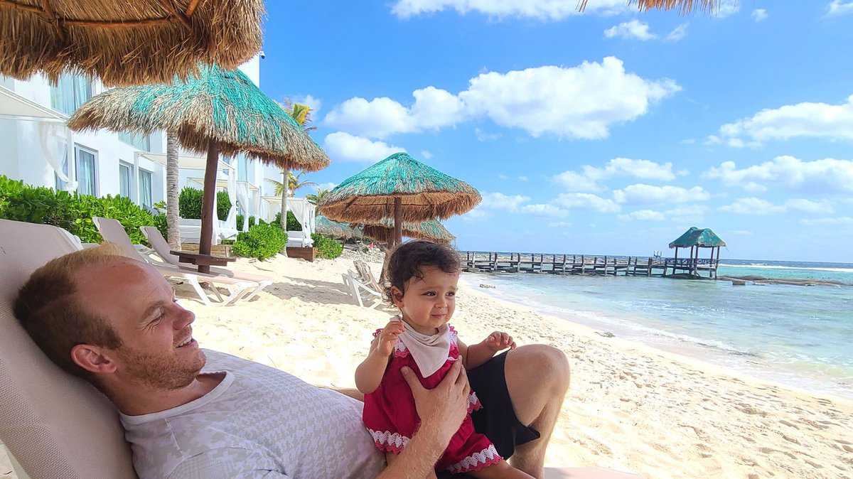 A man and a child relaxing on beach chairs under thatched umbrellas near the shore in Akumal, Quintana Roo, Mexico. In the background are clear blue skies and a wooden pier extending into the sea.