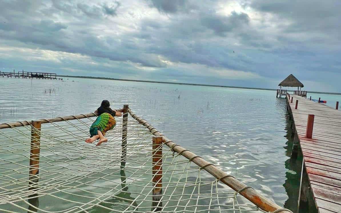 A person lies on a hammock net suspended over the calm waters of Bacalar Lagoon, next to a wooden pier extending into the distance, under a cloudy sky in Bacalar, Quintana Roo, Mexico.
