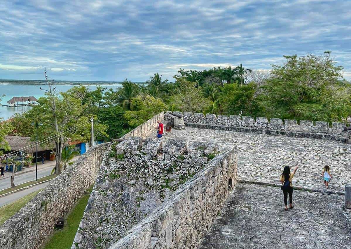 View from Fort San Felipe in Bacalar, Quintana Roo, Mexico, showing an ancient stone wall overlooking lush greenery and the lagoon in the distance under a cloudy sky.