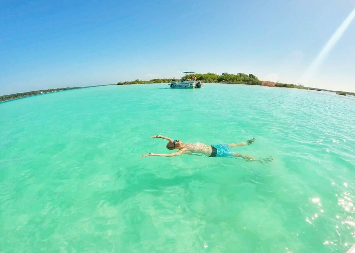 Family Activities in Bacalar Person floating on their back in the clear turquoise waters of Bacalar Lagoon with a boat nearby under a clear blue sky in Bacalar, Quintana Roo, Mexico.