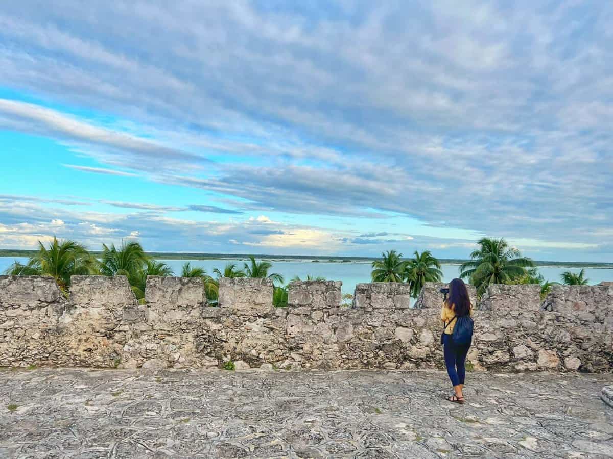 San Felipe Fort - Bacalar A person standing on the stone floor of Fuerte San Felipe in Bacalar, Quintana Roo, Mexico, with a scenic view of palm trees and a lagoon under a partly cloudy sky.