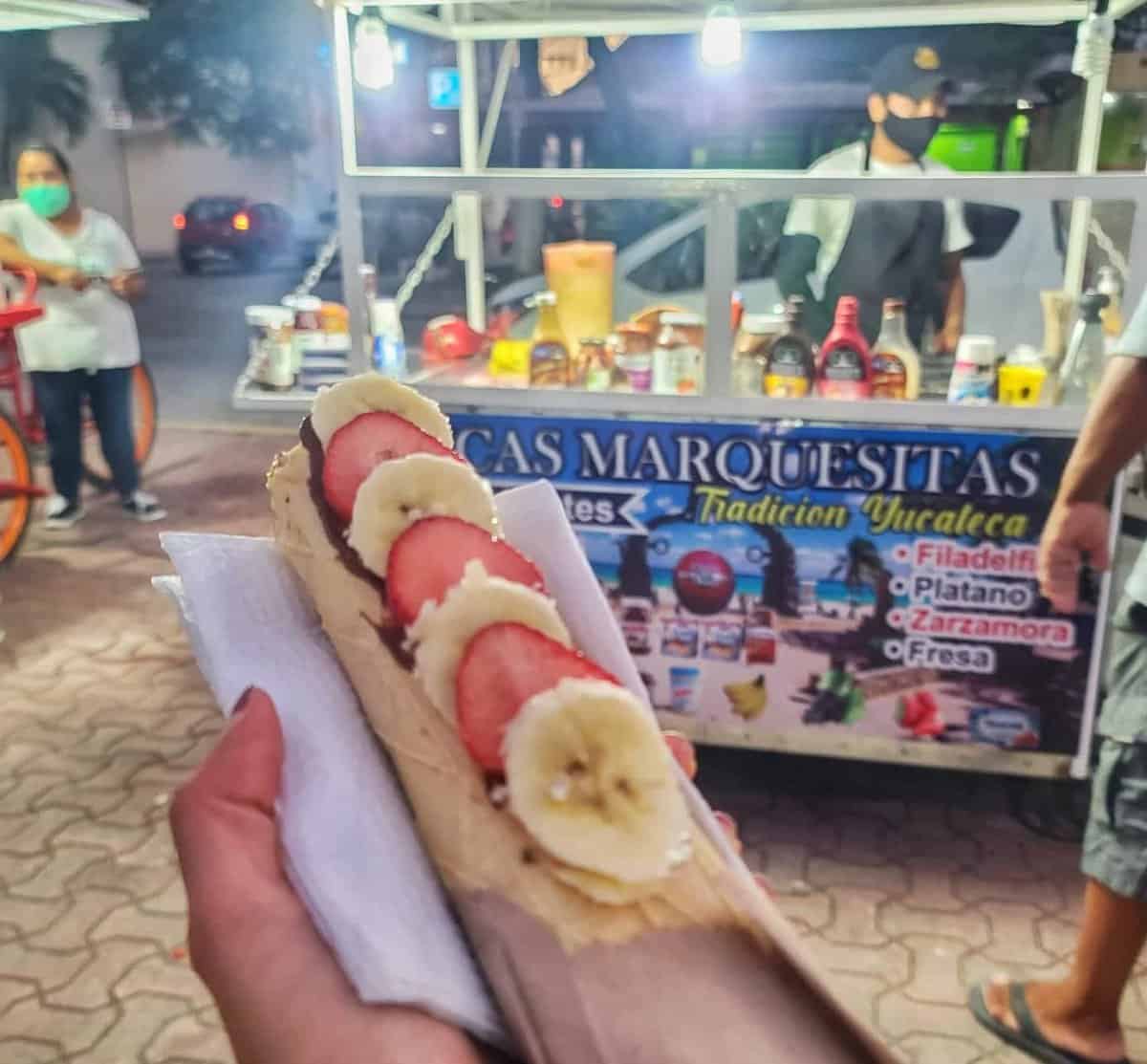 A person holding a filled marquesita with banana slices and strawberries in front of a street food cart in Playa del Carmen, Mexico.