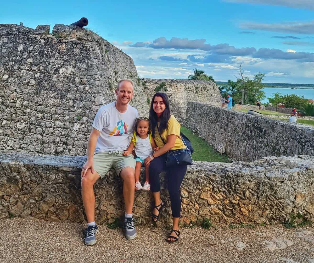 Fort San Felipe - Bacalar What To Do A family of three sits on a stone wall at Fuerte San Felipe in Bacalar, Quintana Roo, Mexico. The historic stone fort and a cannon are visible in the background, along with a clear blue sky.