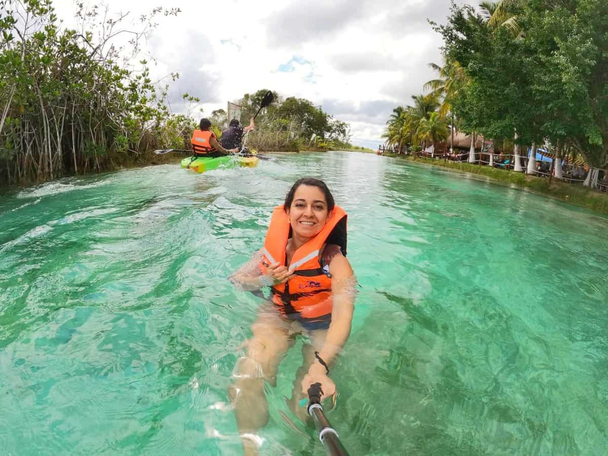Bacalar - Los Rapidos A woman wearing an orange life jacket taking a selfie with a selfie stick while floating in the clear turquoise waters of Los Rapidos in Bacalar, Quintana Roo, Mexico. In the background, a couple is kayaking. The sky is overcast, indicating a rainy day.