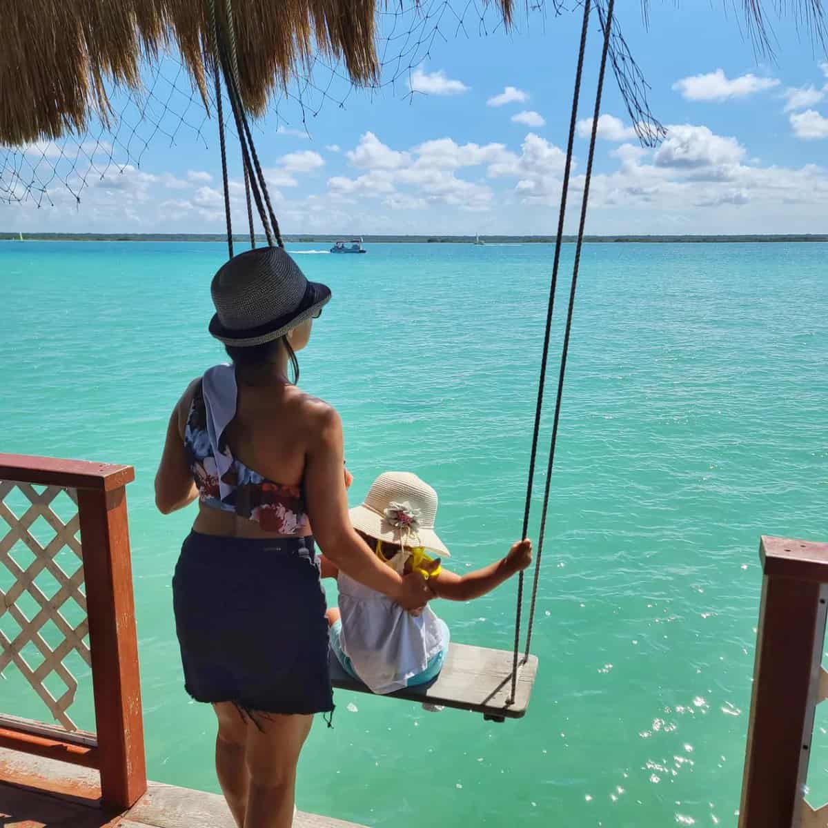 things to do in Bacalar A woman and a child sit on a swing under a thatched roof, overlooking the clear turquoise waters of Bacalar, Quintana Roo, Mexico.