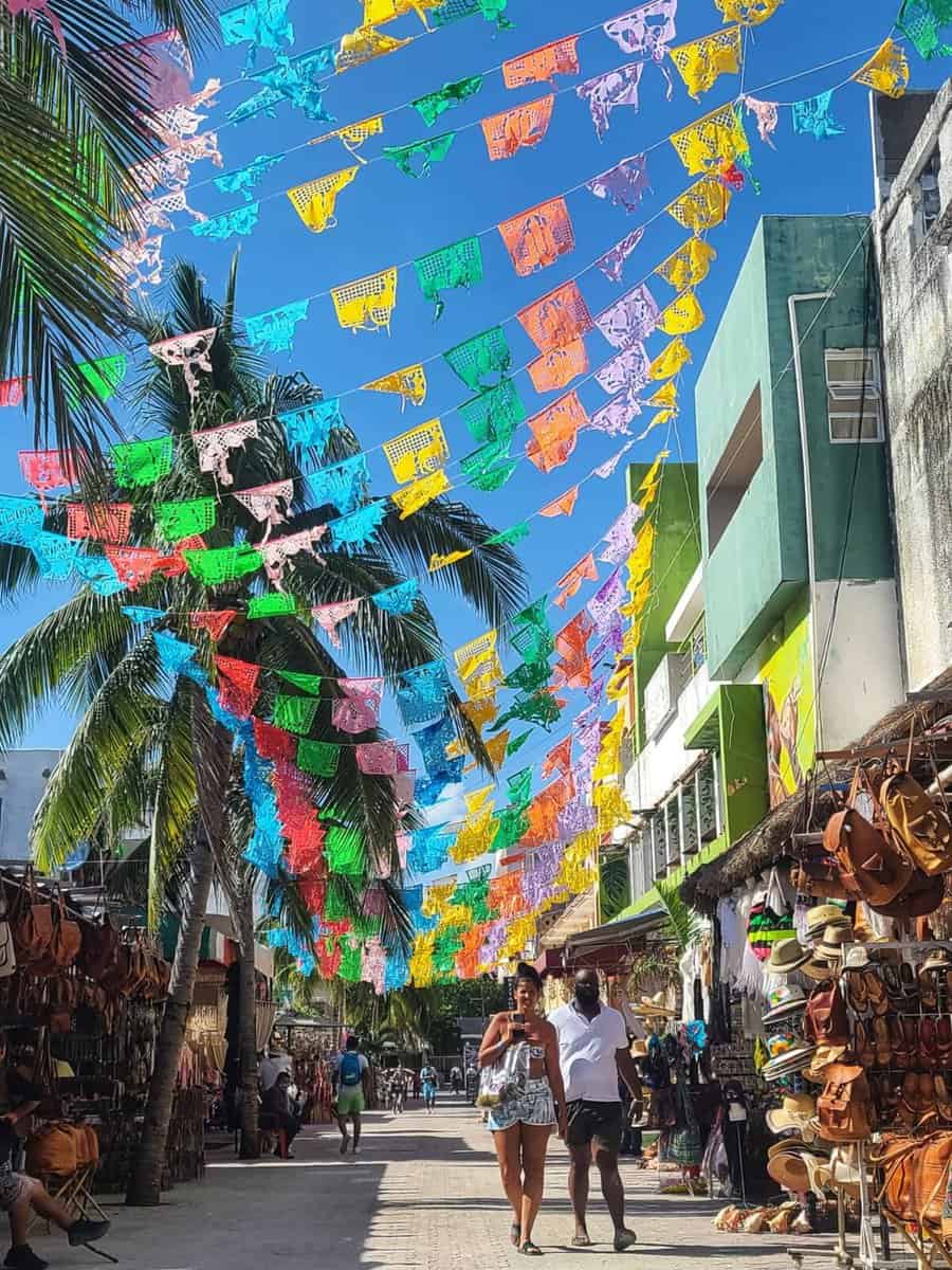 A sunny street view of 5th Avenue in Playa del Carmen, Mexico, with colorful papel picado banners hanging above, palm trees, various shops with leather goods, and people walking.