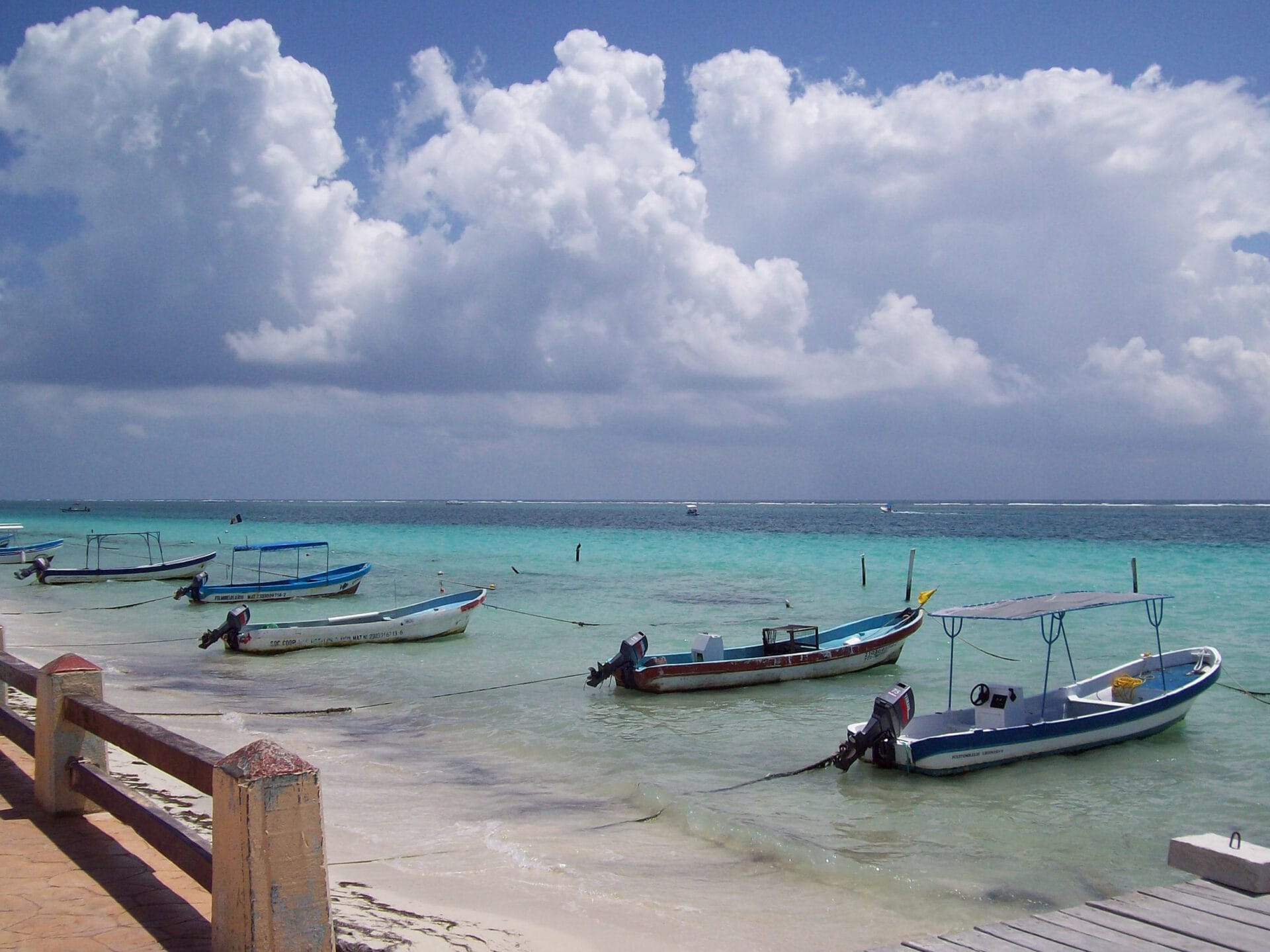 Small boats anchored near the shore in Puerto Morelos, Mexico with a beautiful turquoise sea and partly cloudy sky in the background.
