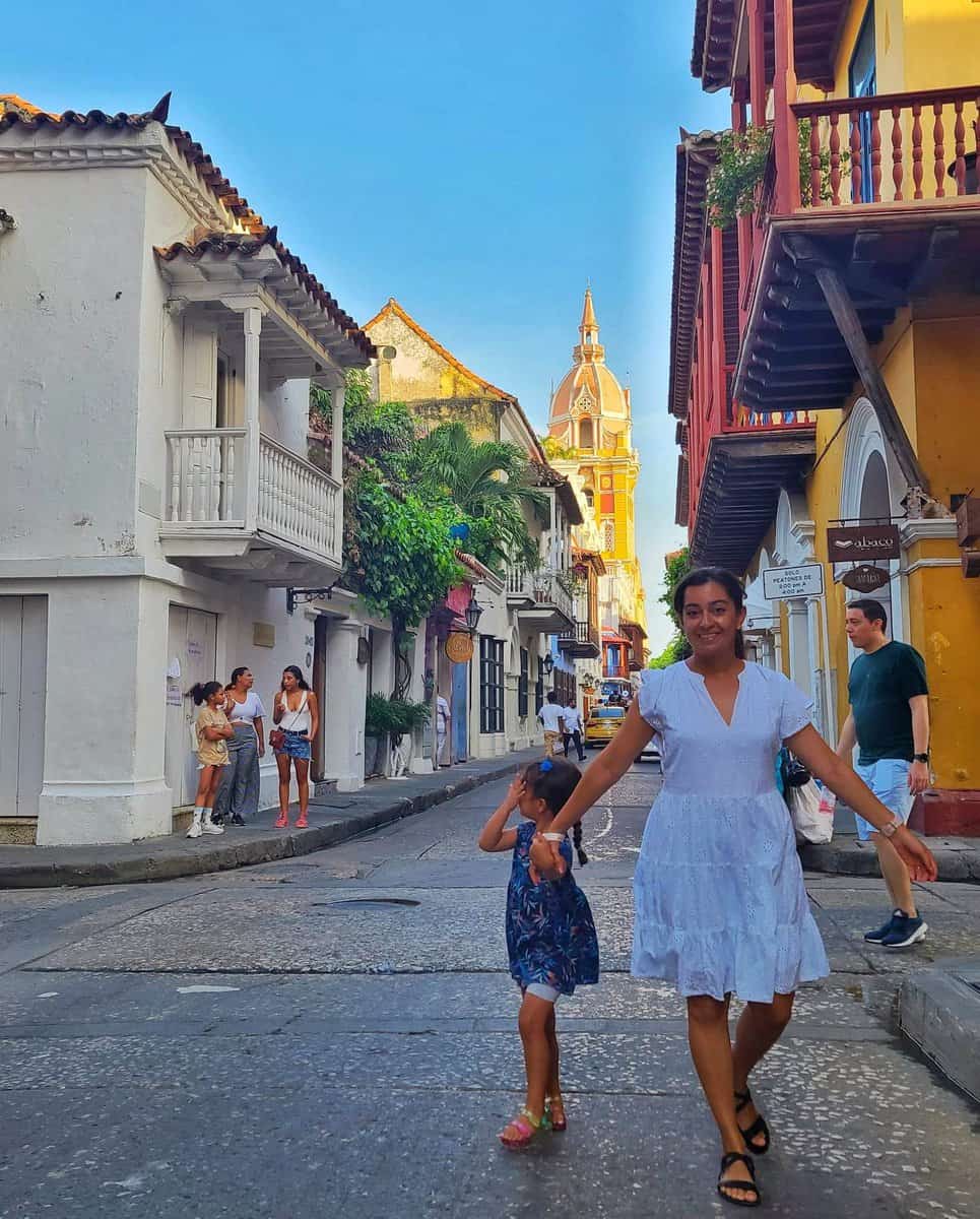A woman and a child walking down a vibrant street in Old Town, Cartagena, Colombia, with colorful colonial architecture and a church tower visible in the background.