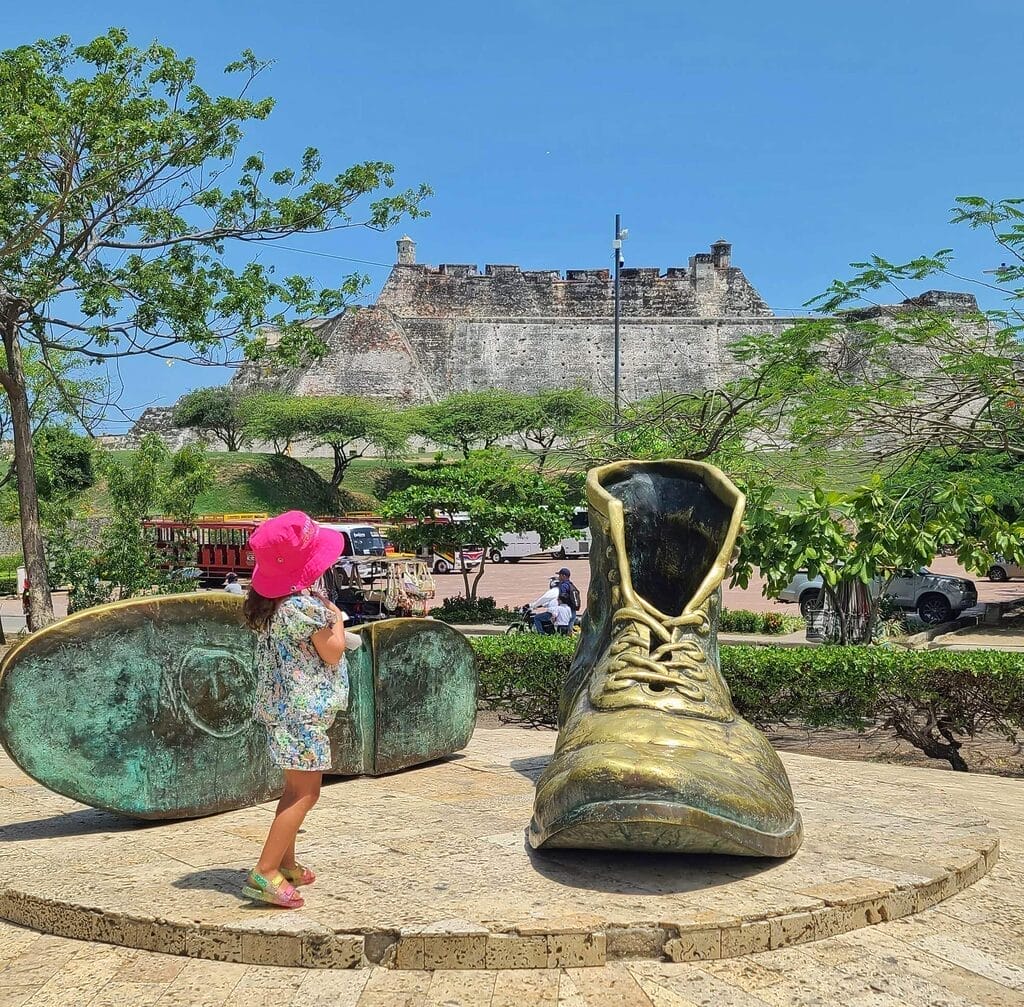 A child in a pink hat stands beside the large bronze Old Shoes Monument near San Felipe Castle in Cartagena, Colombia, with the castle visible in the background.