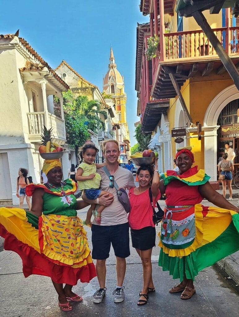A family posing with Palenqueras Fruit Ladies in traditional colorful dresses in Cartagena, Colombia, with a colonial-style street and a church tower in the background.