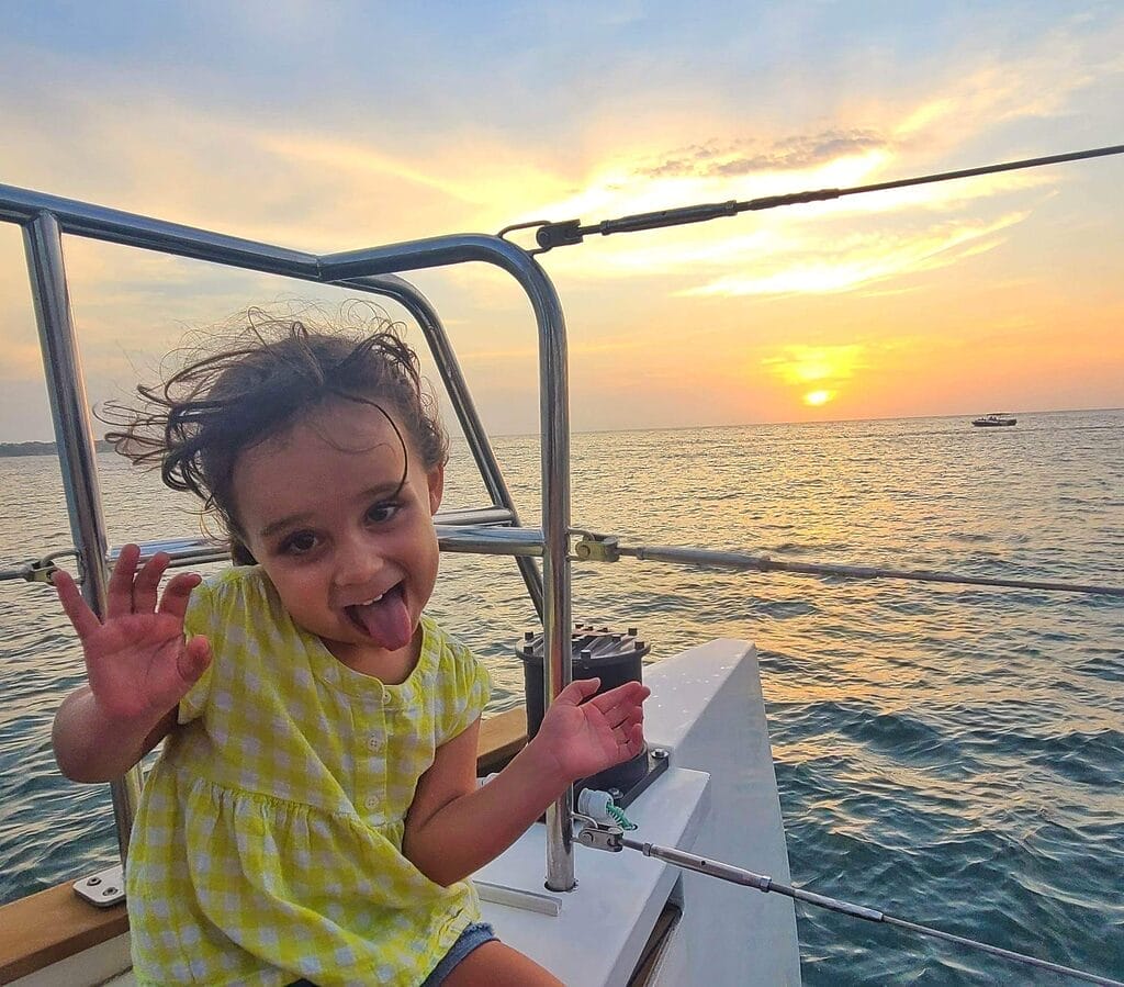 A young child makes a playful face while sitting on a catamaran at sunset, with the ocean and a boat in the background off the coast of Cartagena, Colombia.