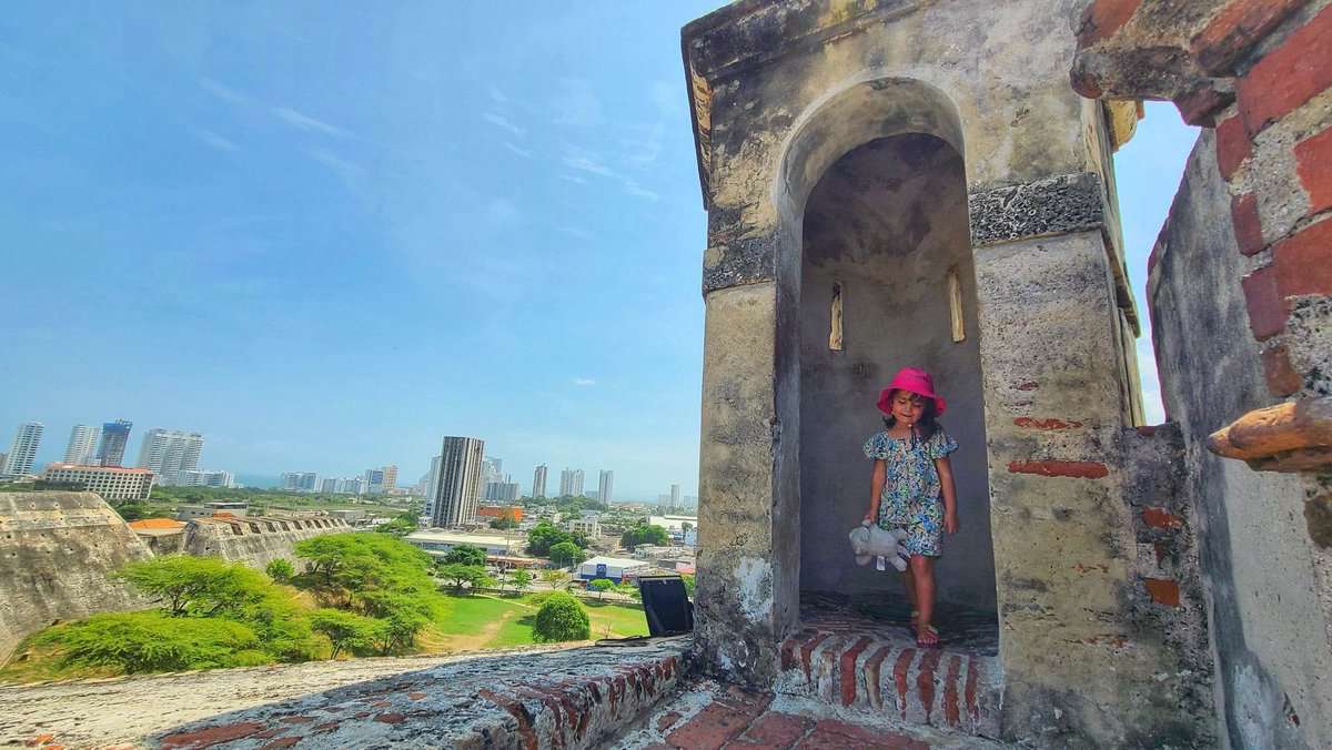 A young girl stands in a stone archway at San Felipe Castle in Cartagena, Colombia, with the city skyline visible in the background.