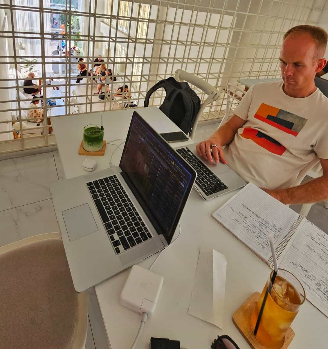 Person working on a laptop at Barisotel in Chiang Mai, Thailand. The table also has a notebook, drinks, a backpack, and charging cables. Several people can be seen conversing in the background.