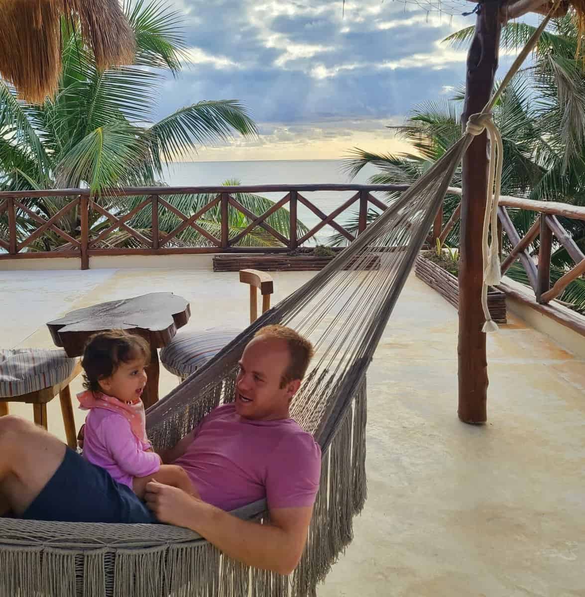 Tulum Mexico compared to Playa Del Carmen A man lounging in a hammock with a young child on a terrace in Tulum, Mexico, with a scenic view of palm trees and the ocean in the background.