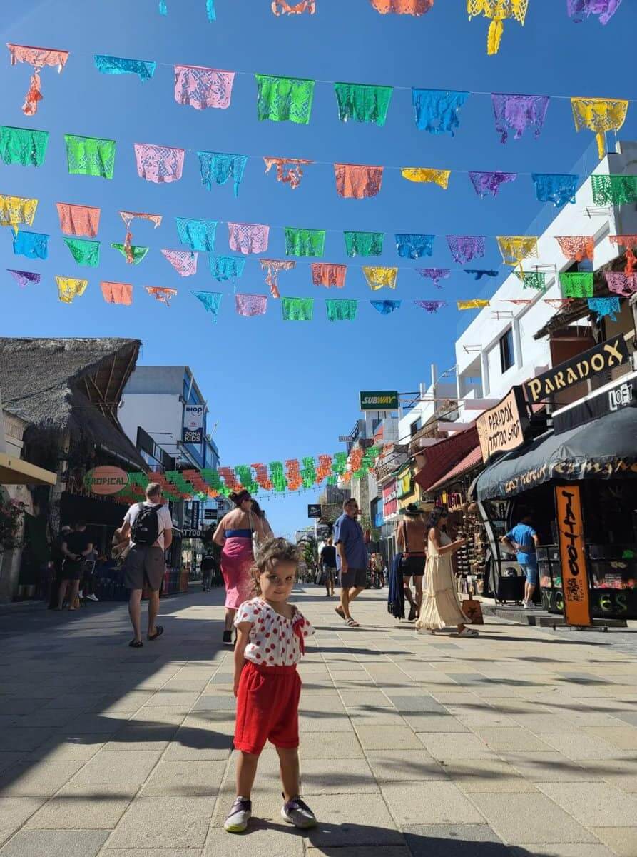 differences between Tulum and Playa Del Carmen Mexico A child stands in the middle of 5th Avenue in Playa del Carmen, Mexico, against a backdrop of colorful banners hanging above the street. People walk past various shops on a sunny day.