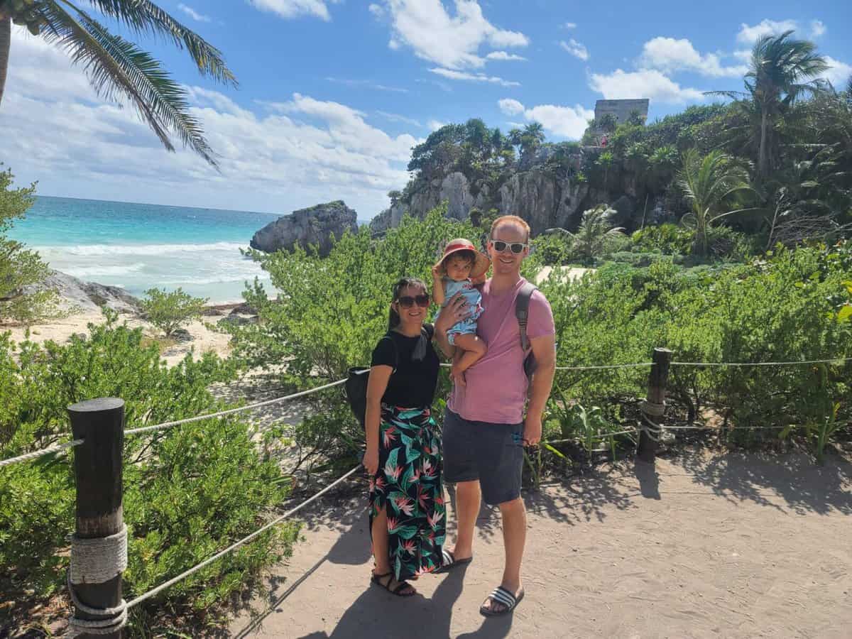Tulum Playa Del Carmen comparison Family standing in front of the Tulum ruins in Mexico with lush greenery, a rocky coastline, and a bright blue sky in the background.