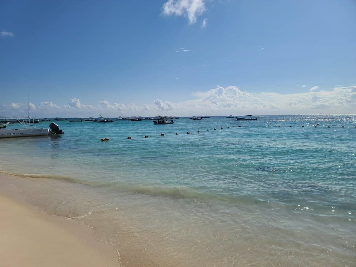 Playa Del Carmen compared to Tulum Boats anchored in the clear blue waters of Tulum, Mexico, with gentle waves lapping at the sandy shore under a bright blue sky.