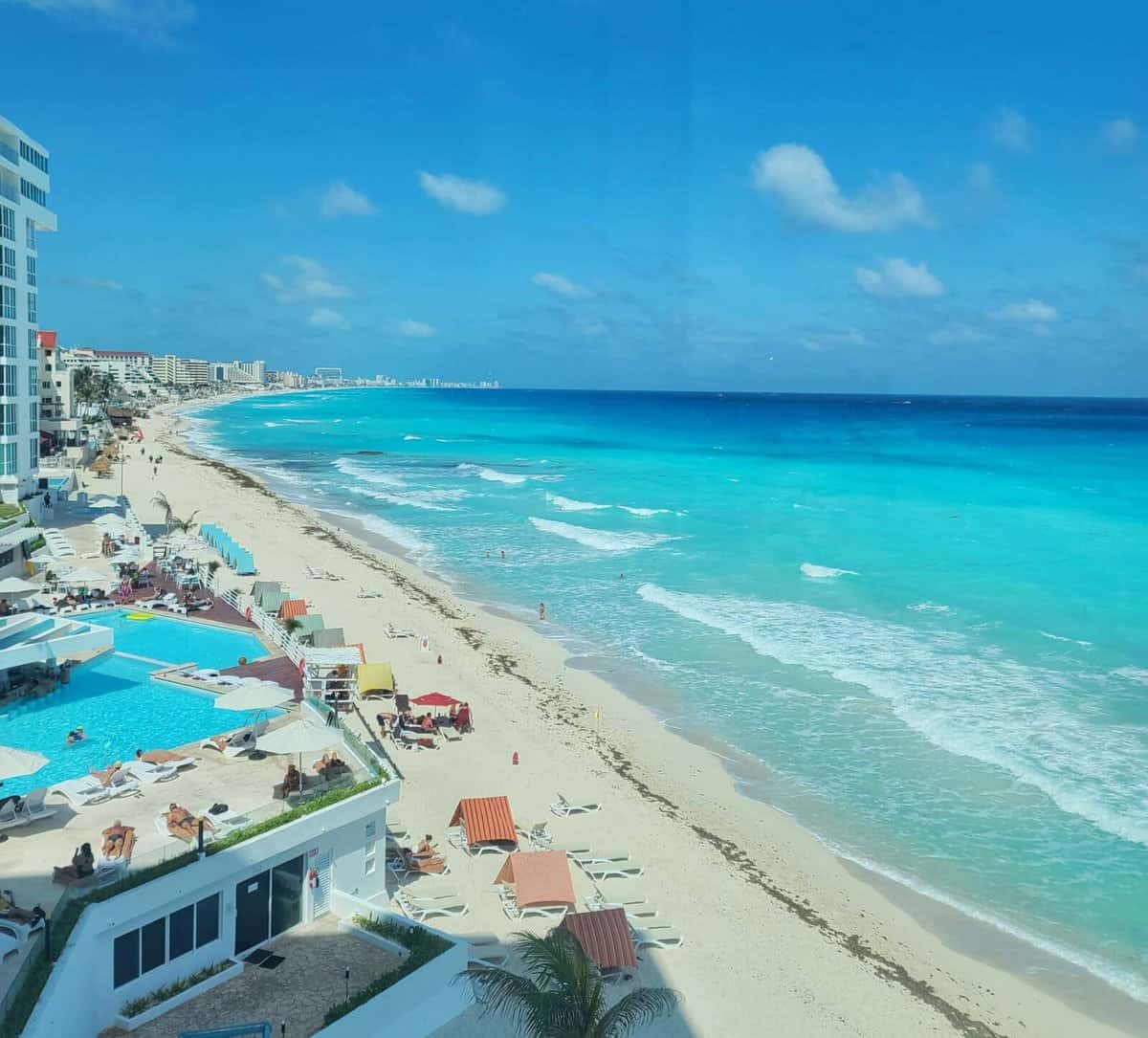 Aerial view of a beautiful beach in Cancun, Mexico, showing crystal-clear turquoise waters, white sandy shore, and lounge chairs and umbrellas along the coastline. 