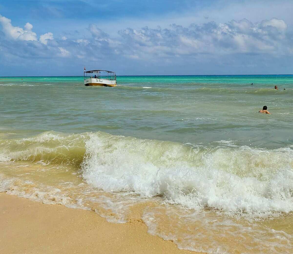 A boat anchored in calm, turquoise waters with gentle waves crashing onto the sandy shoreline at Playa del Carmen, Mexico. Swimmers are enjoying the serene sea.