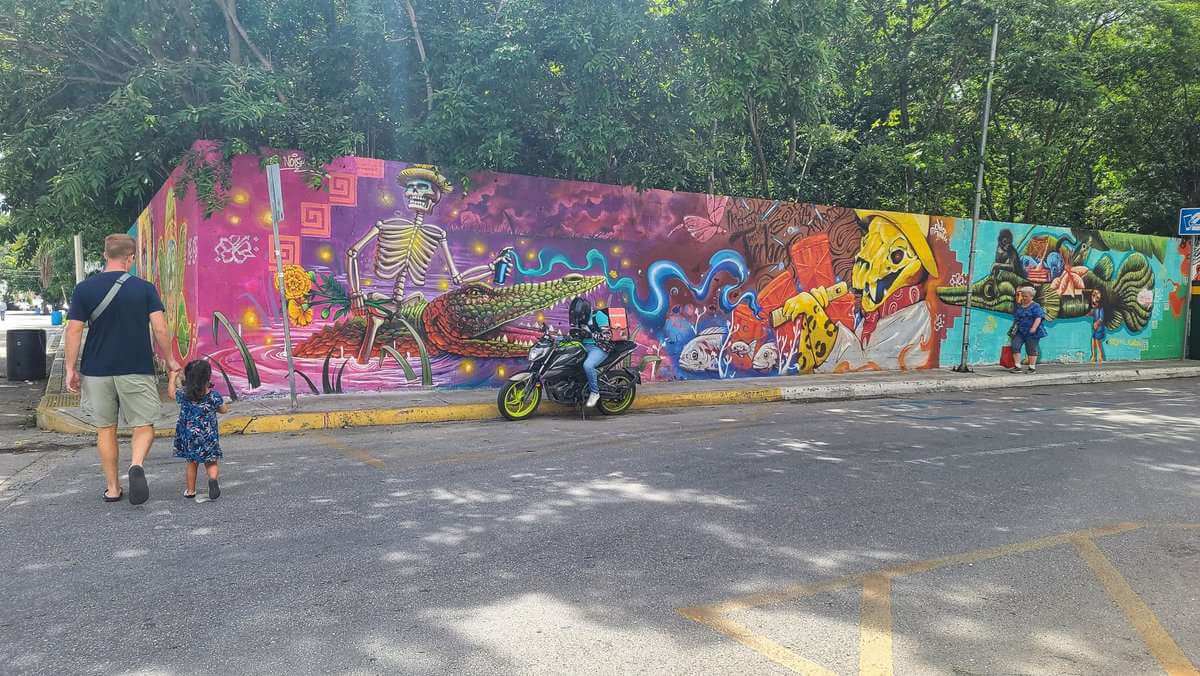 Colorful street mural depicting vibrant characters and scenes, including a skeleton figure riding an alligator, in Playa del Carmen, Mexico. A man and a young girl walk by the mural, while a person on a motorcycle observes the artwork.