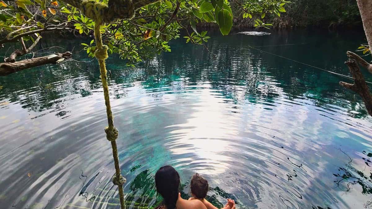 Two people sitting at the edge of the clear, reflective blue waters of Cenote Carwash near Tulum, Mexico, surrounded by lush green foliage.