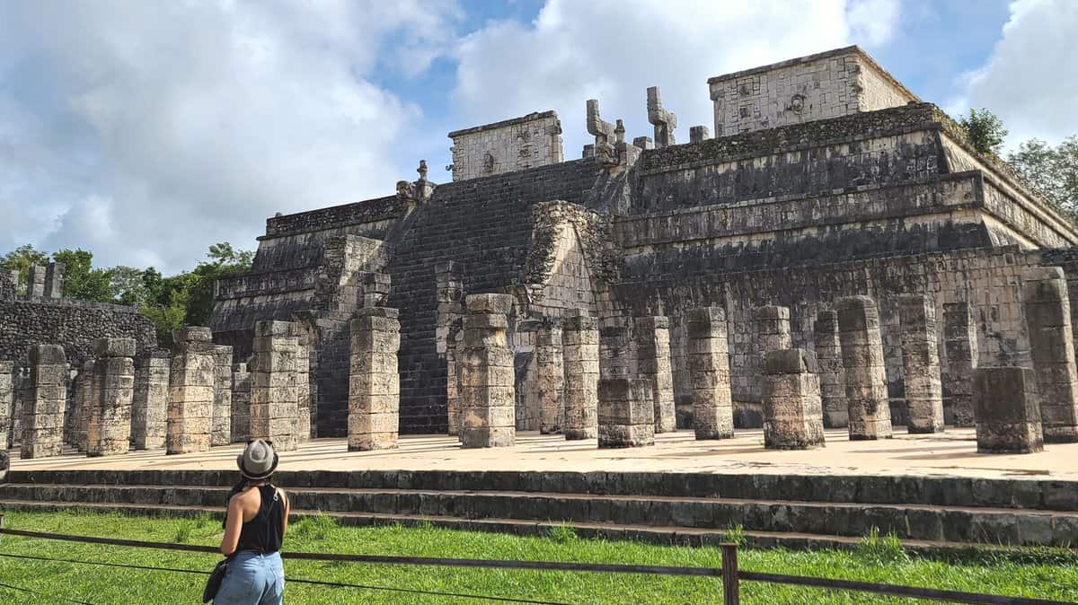 A visitor in a hat stands in front of the Temple of the Warriors, an ancient Mayan structure at Chichen Itza, Mexico, on a sunny day with scattered clouds in the sky.