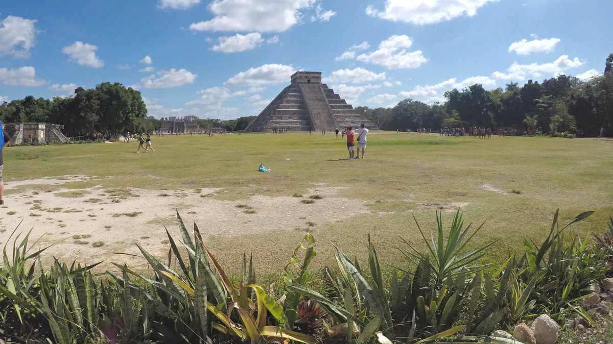 Wide view of the Pyramid of Kukulkan at Chichen Itza, with people exploring the surrounding grounds under a bright, partially cloudy sky.