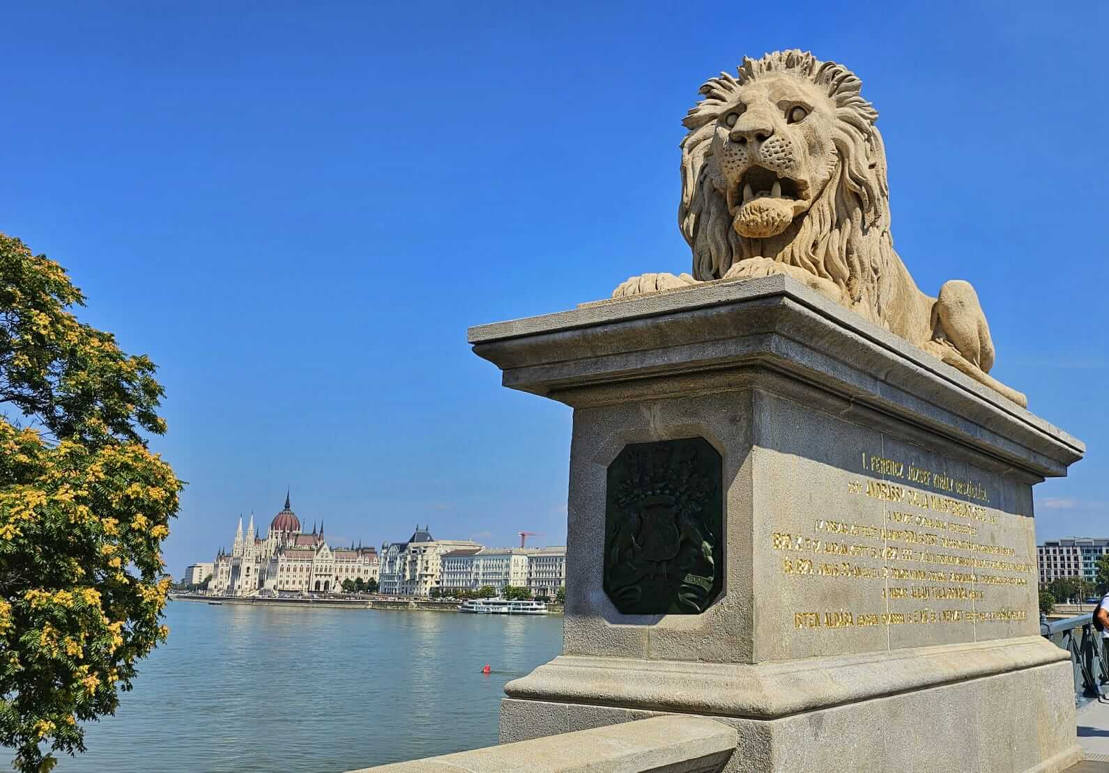 A stone lion statue on the Széchenyi Chain Bridge in Budapest, Hungary, with the Hungarian Parliament Building visible across the Danube River.