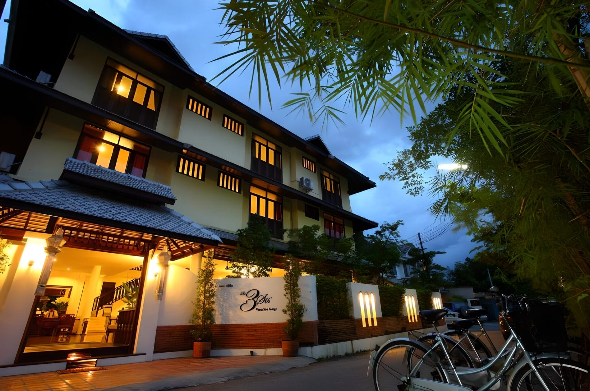 Front facade of 3sis Hotel in Chiang Mai, Thailand, at dusk with lit windows and bicycles parked outside.