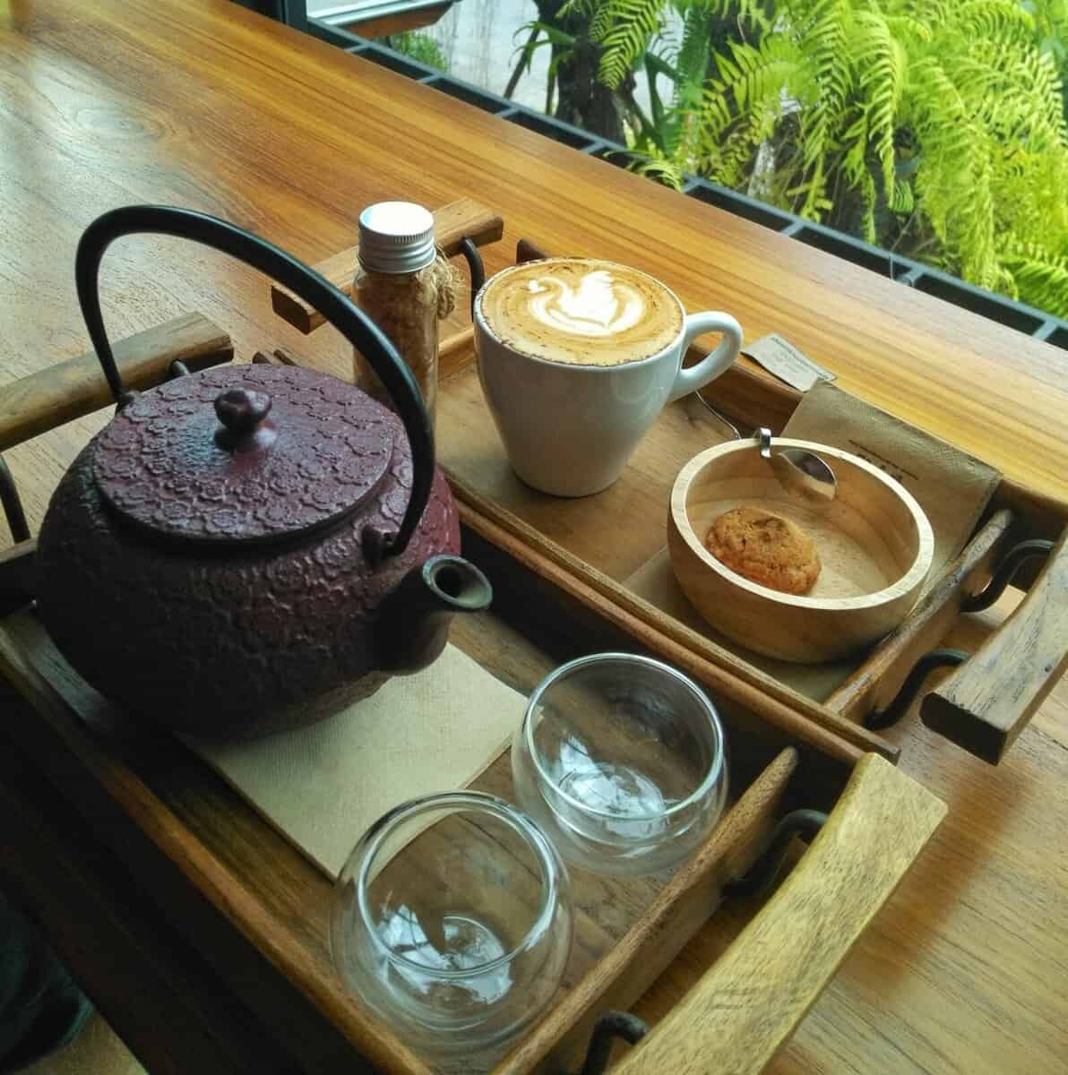 A cozy cafe setup in Chiang Mai, Thailand, featuring a rustic teapot, two glass cups, a cup of foamy coffee, a jar of brown sugar, and a small cookie on a wooden tray.