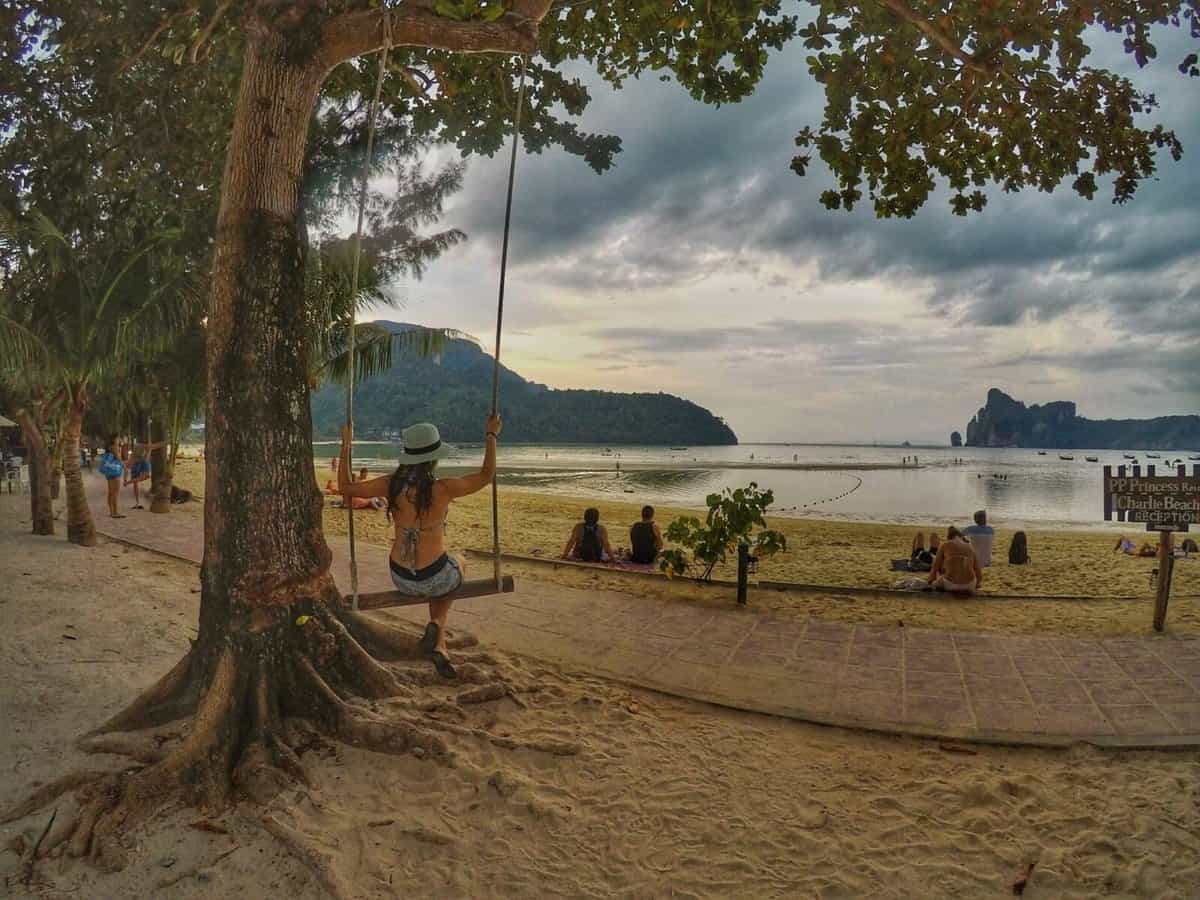 A woman on a swing hanging from a tree on a sandy beach in Koh Phi Phi, Thailand. In the background, people enjoy the beach and the scenic view of the mountains and the sea.