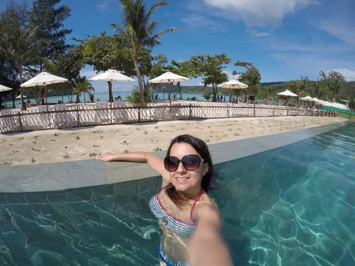 Woman in sunglasses relaxing in a swimming pool with a beach and umbrellas in the background at PP Charlie Beach Resort on Koh Phi Phi, Thailand.