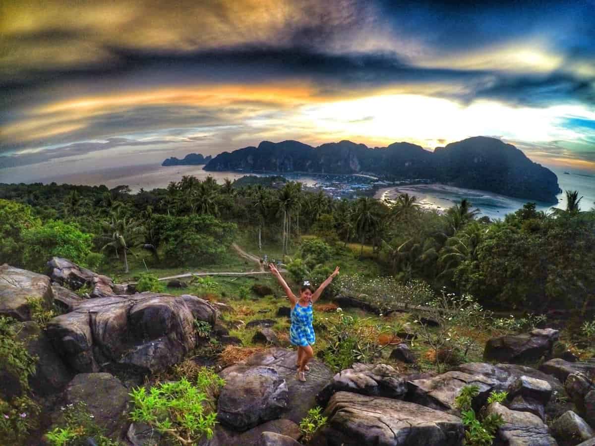 Person posing joyfully on rocks at Koh Phi Phi Viewpoint in Thailand with a stunning sunset and panoramic view of lush greenery and distant mountains in the background.