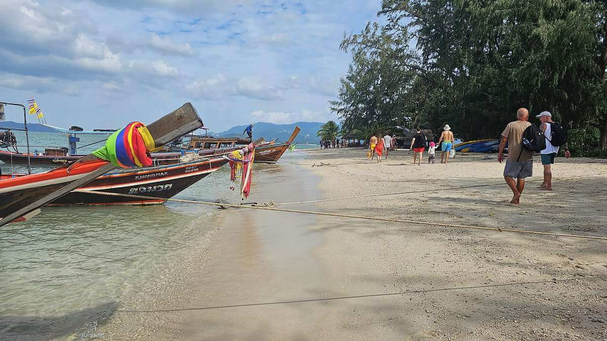 Tourists walking on the sandy beach of Pig Island (Koh Madsum) in Thailand with traditional long-tail boats moored along the shore.
