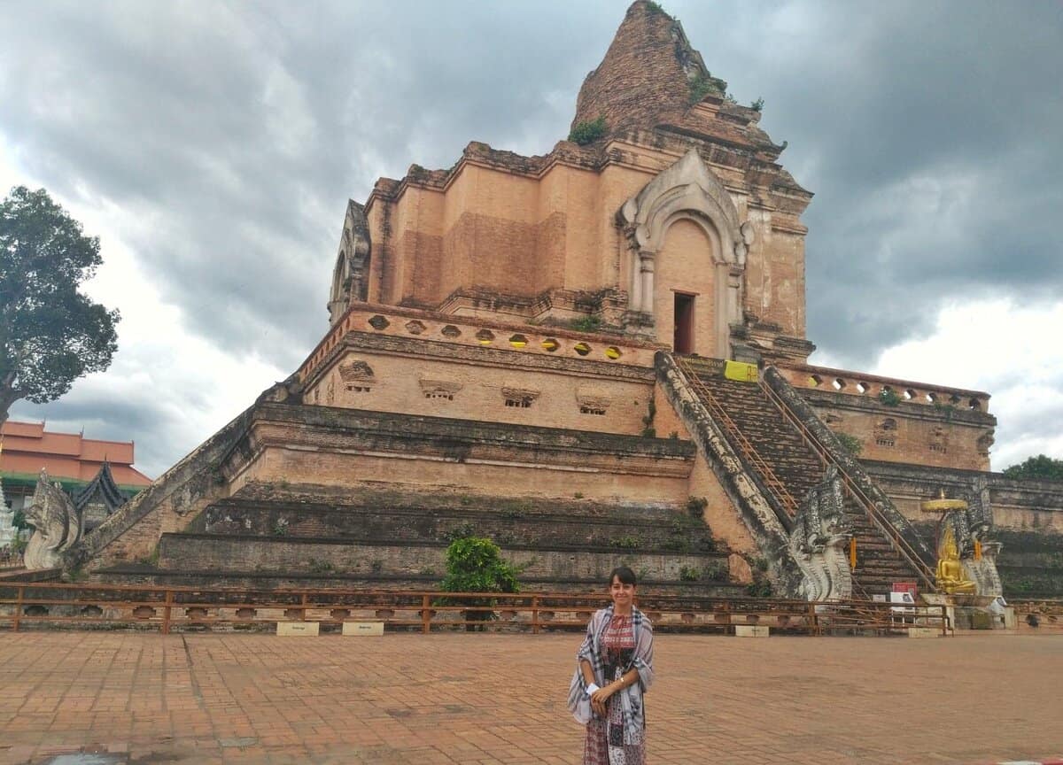 A person standing in front of the ancient Wat Chedi Luang in Chiang Mai, Thailand. The large brick temple is partially ruined with steep steps leading to a doorway, and the sky is overcast with clouds.