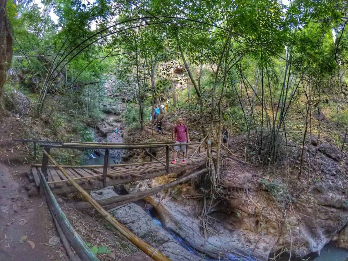 A wooden bridge over a small stream surrounded by lush greenery and bamboo trees in Pai, Thailand. A person is standing on the bridge, while others explore the area in the background.