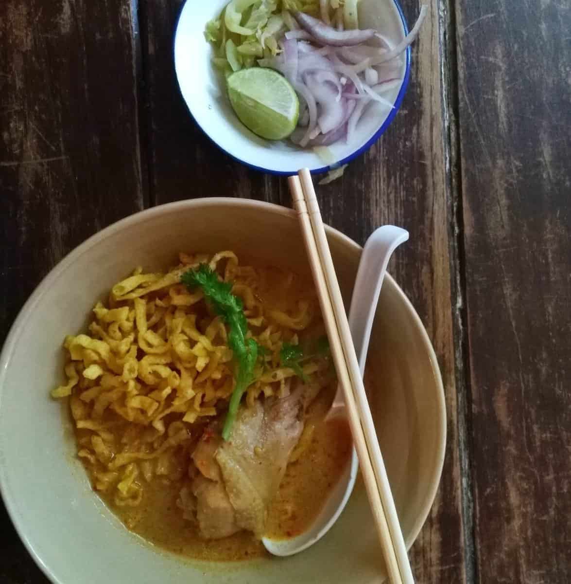 A bowl of traditional Khao Soi, a popular Northern Thai curry noodle soup, on a rustic wooden table in Chiang Mai, Thailand. It is garnished with a sprig of cilantro and served with chopsticks and a ceramic spoon. A small dish of sliced shallots, pickled mustard leaves, and a lime wedge accompanies the dish.