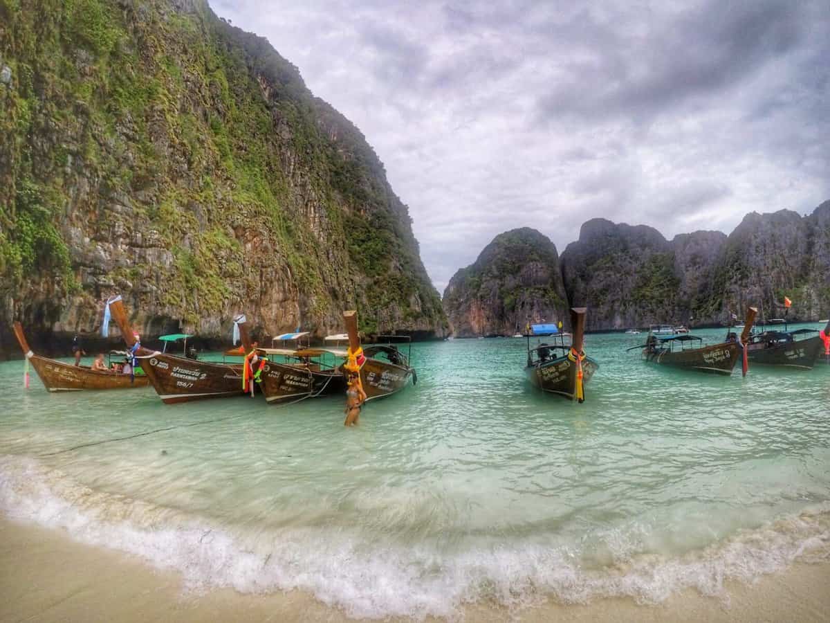 Traditional long-tail boats anchored in the clear turquoise waters of Maya Bay, Thailand, with towering green cliffs in the background.